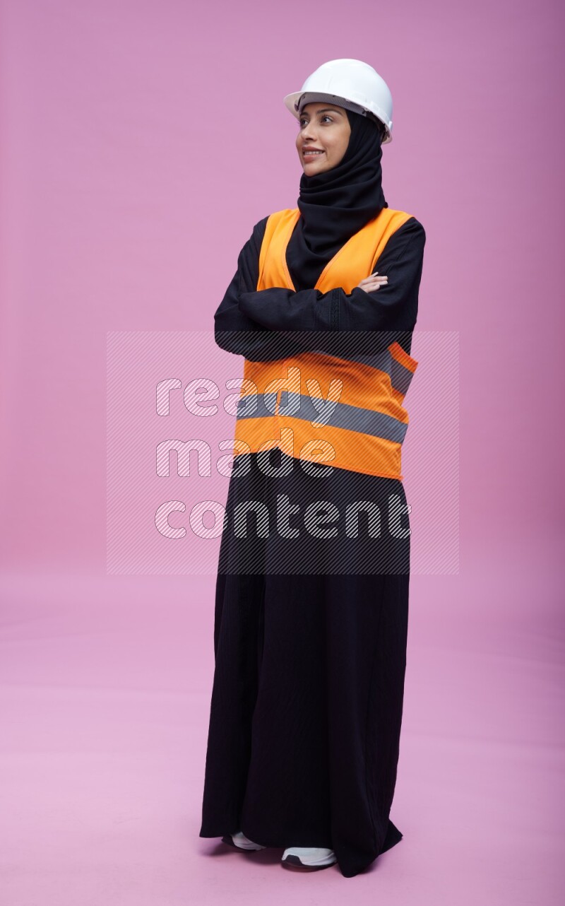 Saudi woman wearing Abaya with engineer vest and helmet standing with crossed arms on pink background