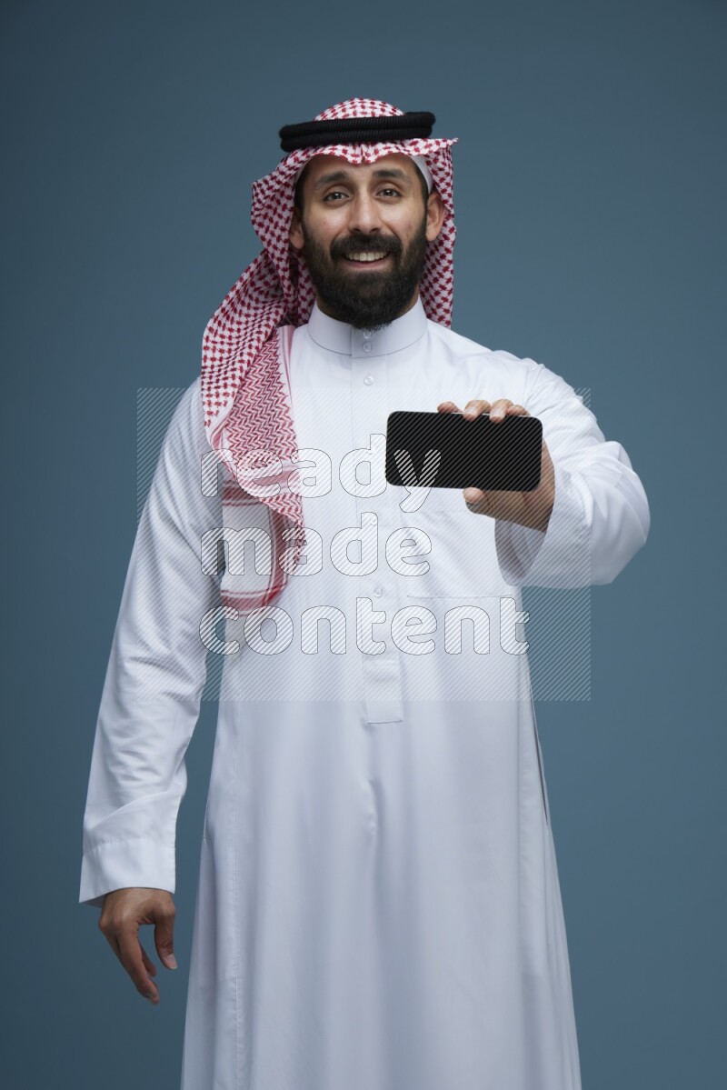 A man showing his phone Screen in a blue background wearing Saudi Thob with Shomag