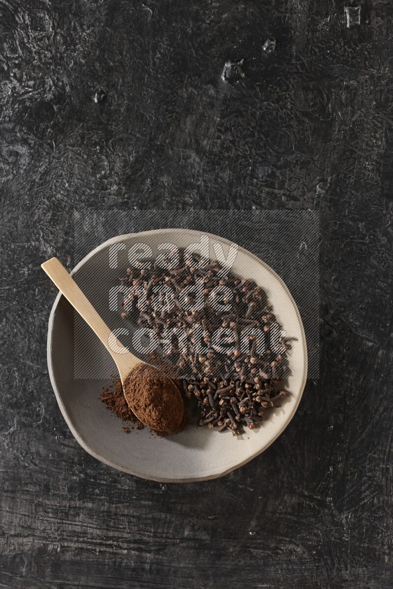 A Pottery plate full of cloves and a wooden spoon full of cloves powder on it on a textured black background