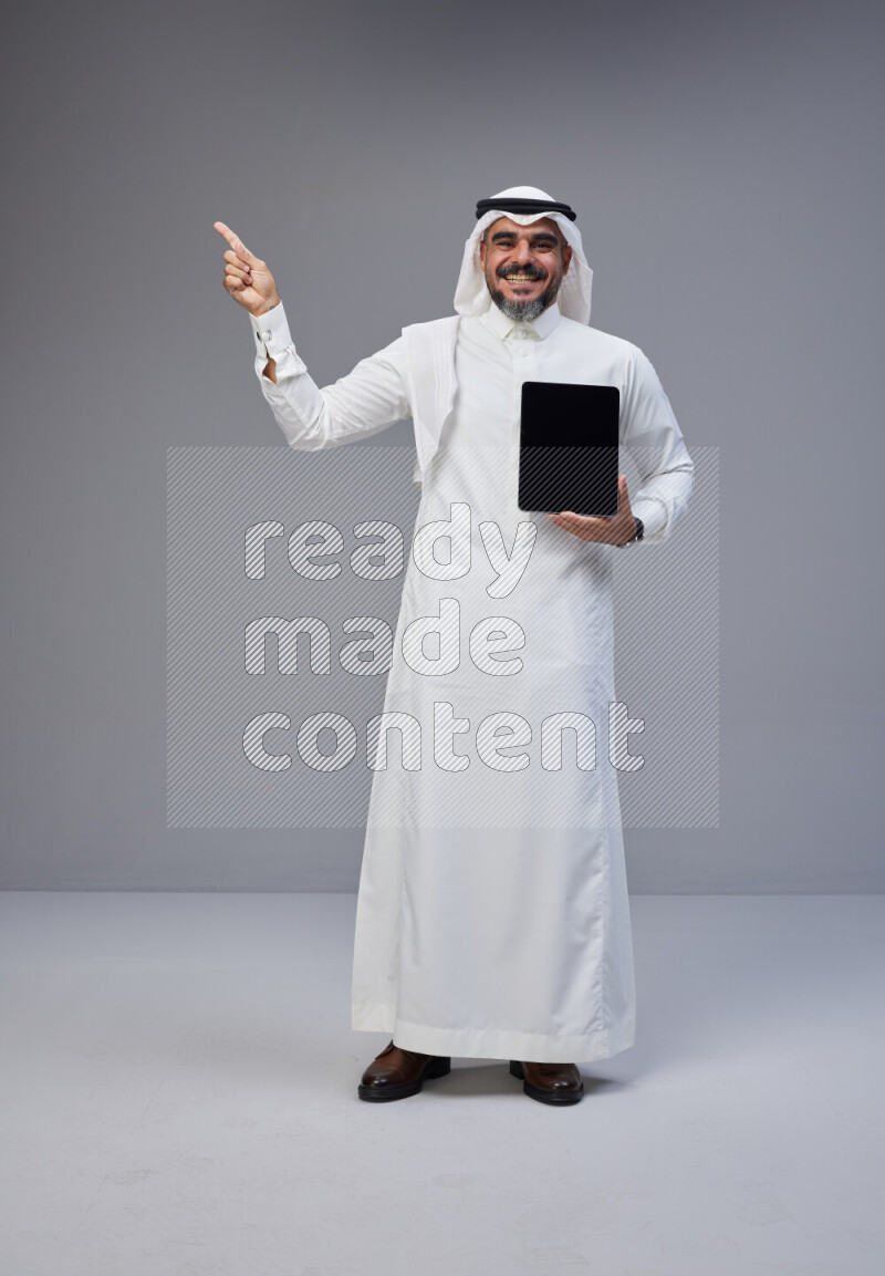 Saudi man Wearing Thob and white Shomag standing showing tablet to camera on Gray background