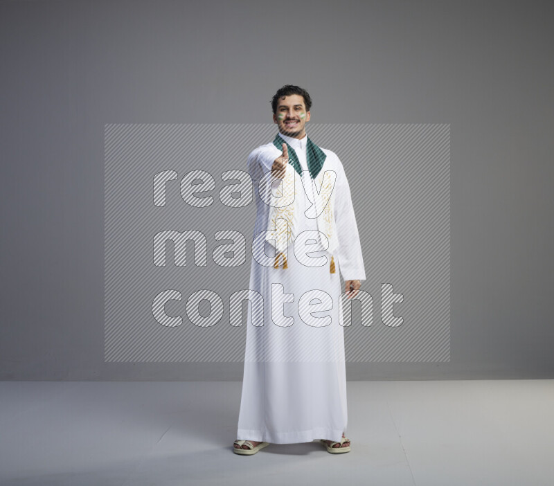 A Saudi man standing wearing thob and Saudi flag scarf with face painting on gray background