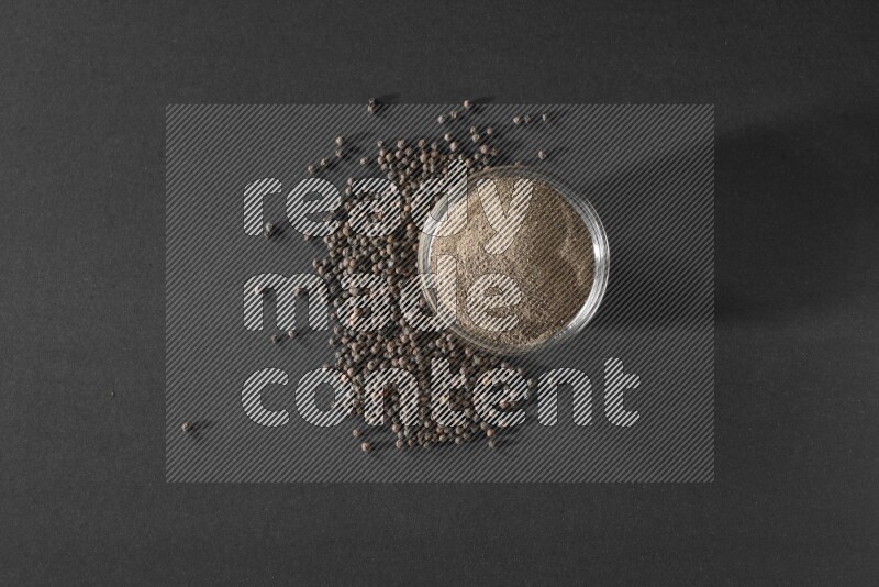 A glass bowl full of powder black pepper powder and black pepper beads on the floor on black flooring