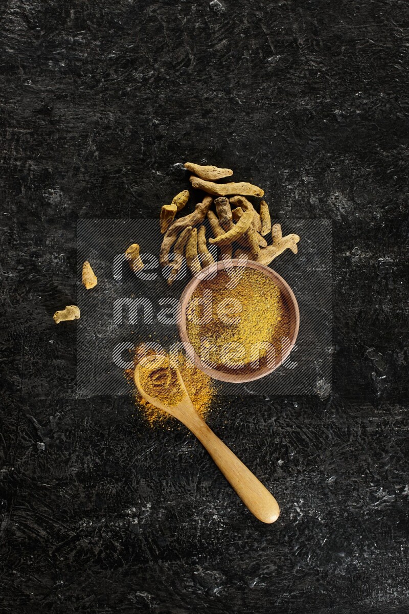A wooden bowl and a wooden spoon full of turmeric powder with dried turmeric fingers on textured black flooring