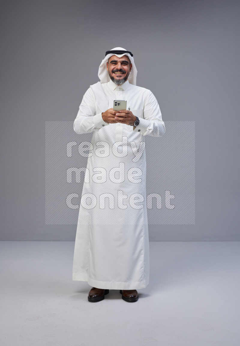 Saudi man Wearing Thob and white Shomag standing texting on phone on Gray background