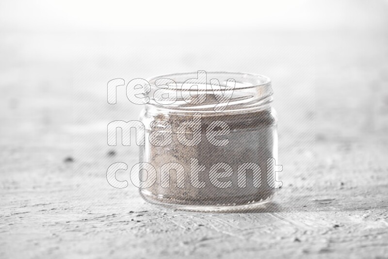A glass jar full of black pepper powder on a textured white flooring