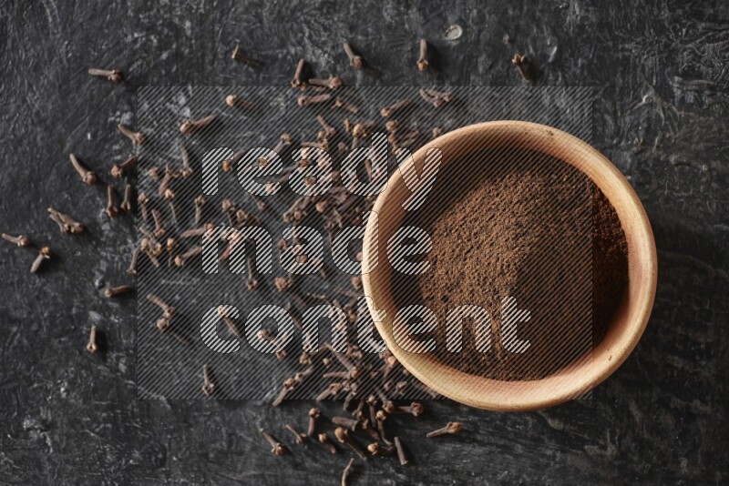 A wooden bowl full of cloves powder on a textured black flooring