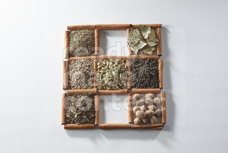 9 squares of cinnamon sticks full of cardamom in the middle surrounded by nutmeg, salt, dried basil, white pepper, cumin, allspice, bay laurel leaves and black pepper on white flooring