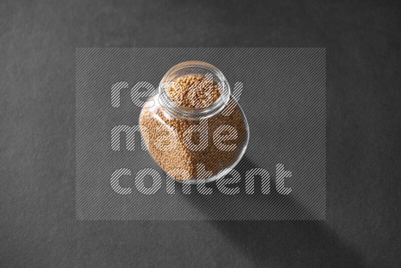 A glass spice jar full of mustard seeds on black flooring