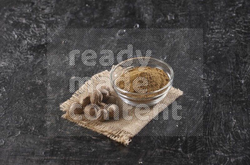 A glass bowl full of nutmeg powder with whole seeds beside it on burlap fabric on a textured black flooring