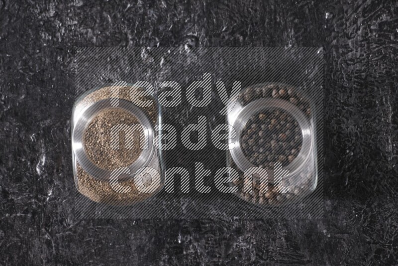2 glass spice jars full of black pepper powder and black pepper beads on textured black flooring