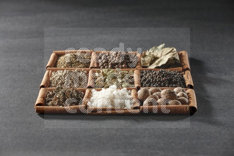 9 مربعات من أعواد القرفة ممتلئة بالهيل في المنتصف محاطة بجوزة الطيب والفلفل الأسود وورق الغار والبهارات والكمون والفلفل الأبيض والريحان المجفف والملح على أرضية سوداء