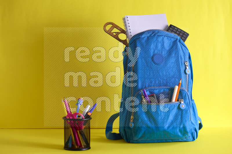A school bag with assorted school supplies in and beside it on yellow background