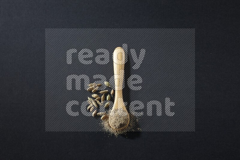 ملعقة خشبية ممتلئة ببودرة الهيل وحبوب الهيل بجانبها على خلفية سوداء