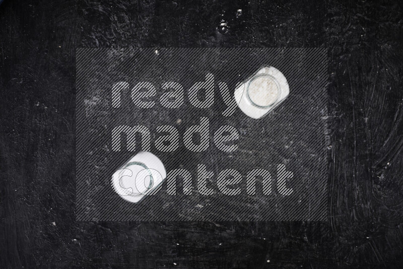 2 وعاء زجاجي احدهمها ممتلئ بملح الطعام الأبيض والأخر بملح البحر الخشن على خلفية سوداء