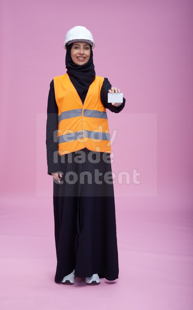 Saudi woman wearing Abaya with engineer vest and helmet standing holding ATM card on pink background