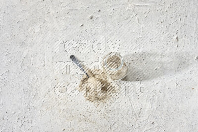 Herbal Glass jar full of white pepper powder and metal spoon full of powder on textured white flooring
