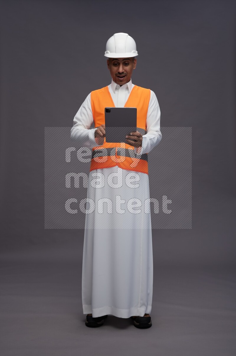 Saudi man wearing thob with engineer vest standing working on tablet on gray background