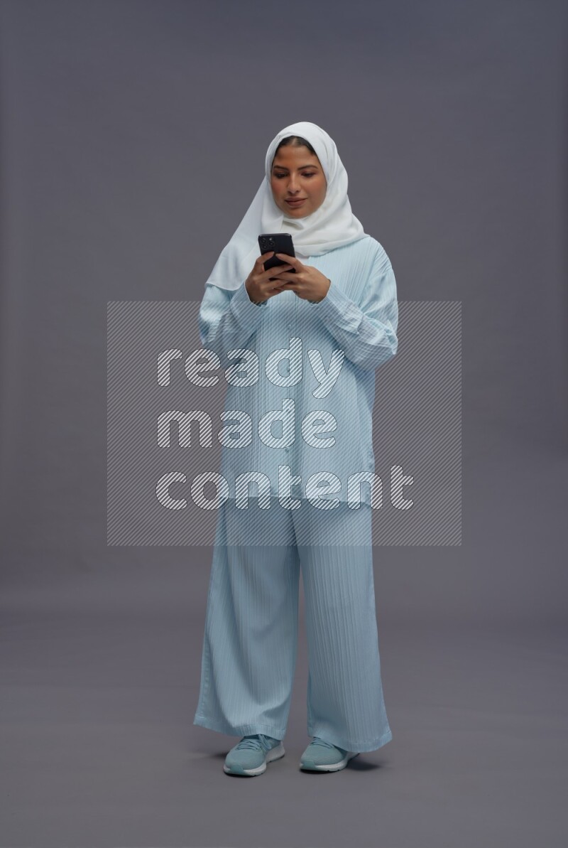 Saudi woman wearing hijab clothes standing texting on phone on gray background