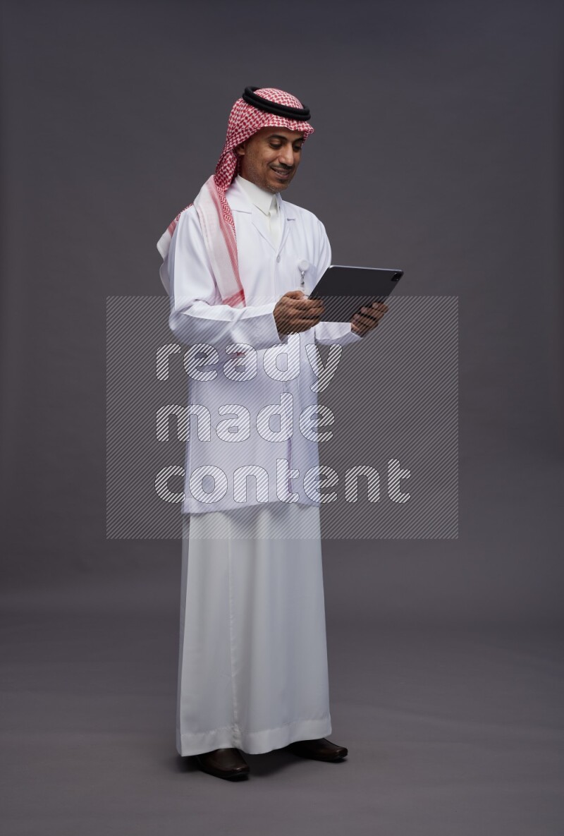 Saudi man wearing thob with lab coat and shomag with pocket employee badge standing working on tablet on gray background