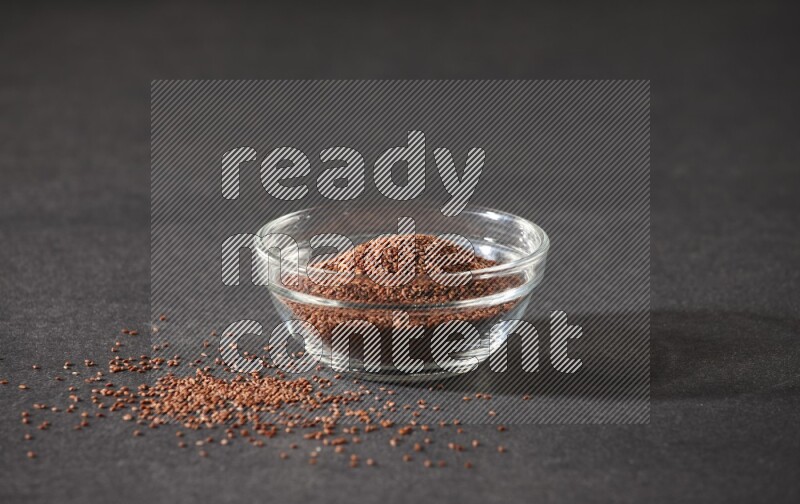 A glass bowl full of garden cress seeds surrounded by seeds on a black flooring