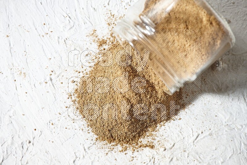 A flipped glass jar full of cumin powder and powder spilled out on textured white flooring