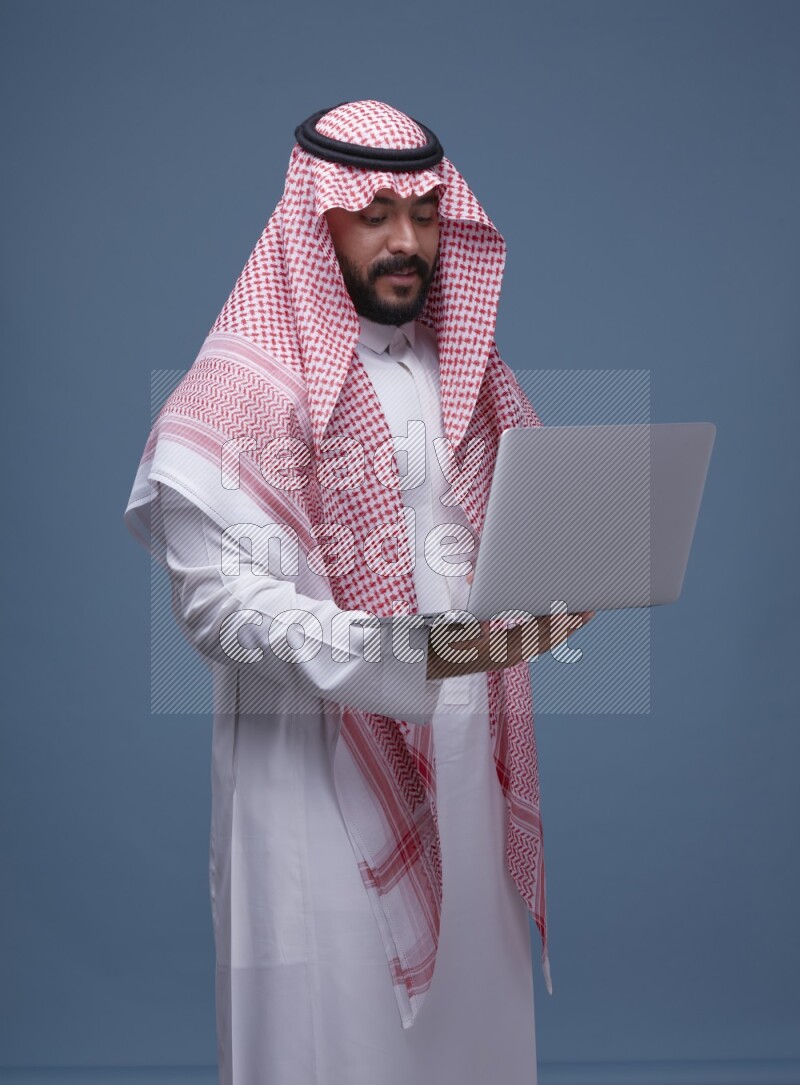 A man standing  with a laptop on Blue Background wearing Saudi Thob and Shomag