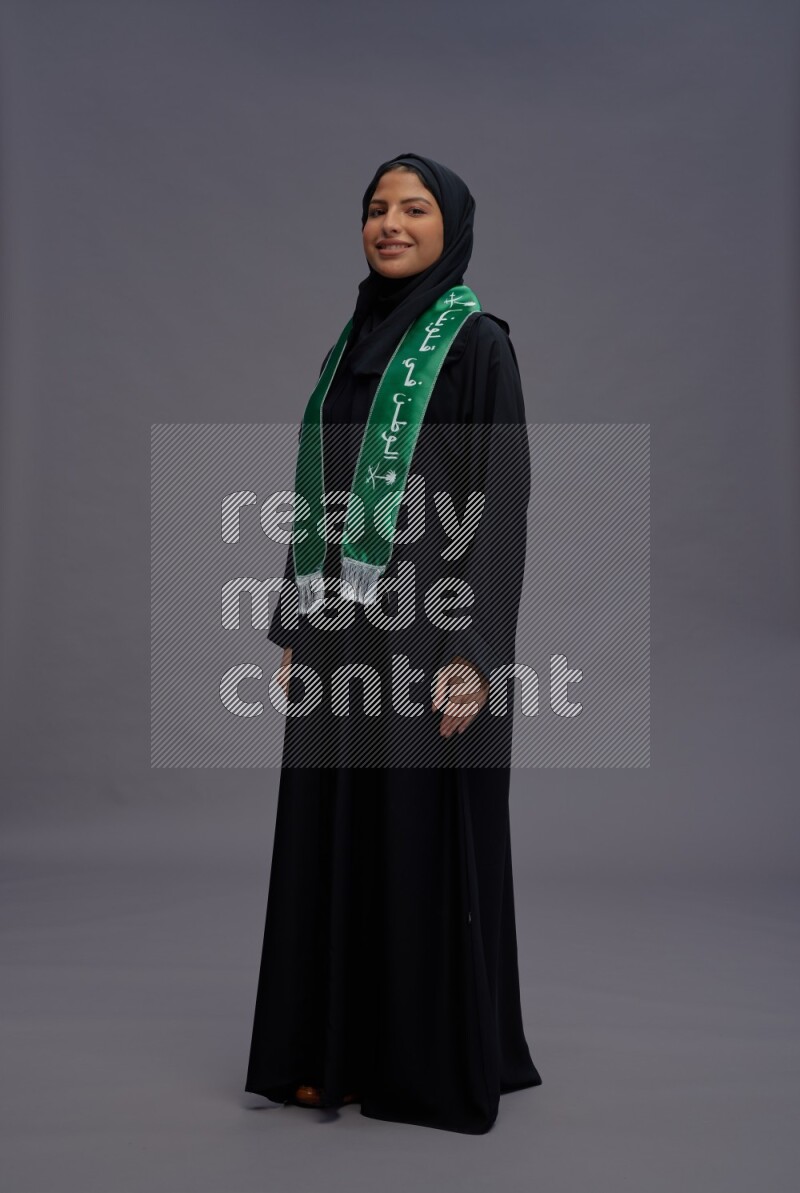 Saudi woman wearing Abaya standing holding Saudi flag on gray background