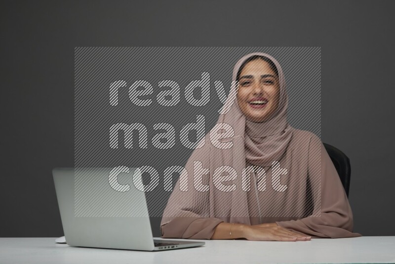 A Saudi woman Setting on her desk on a Gray Background wearing Brown Abaya with Hijab