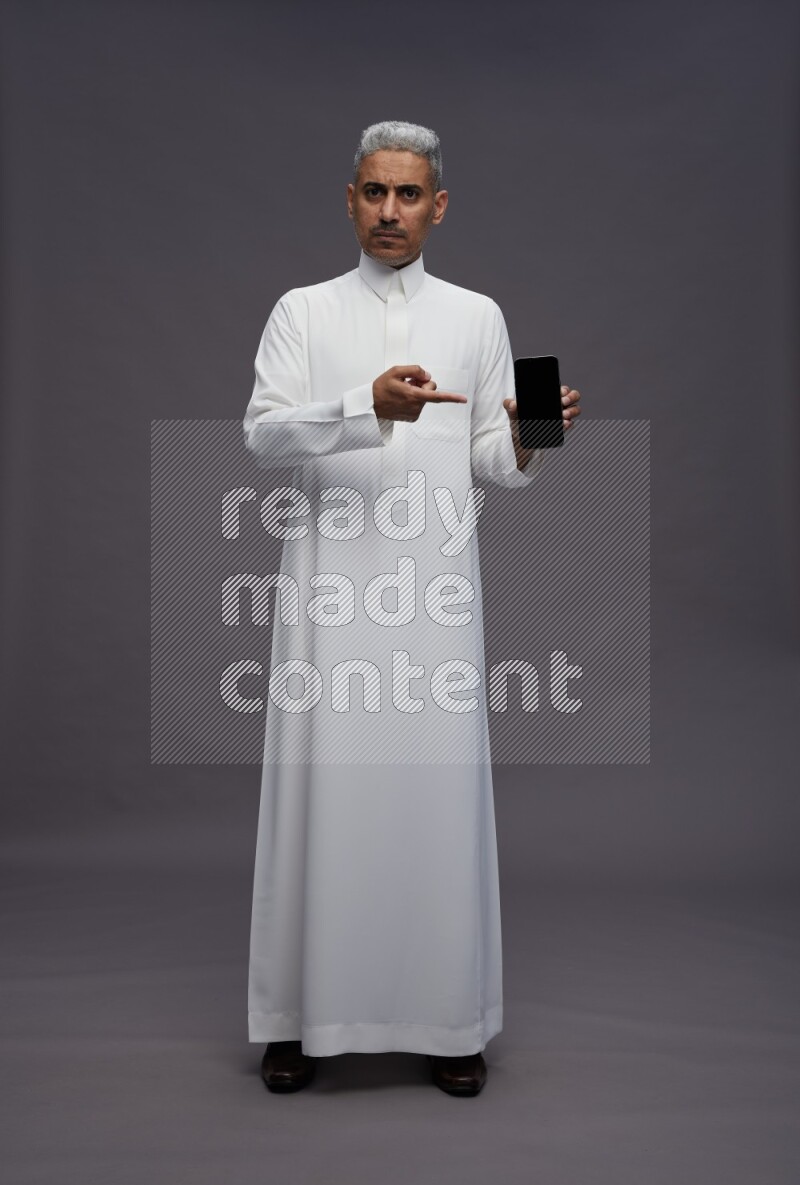 Saudi man wearing thob standing showing phone to camera on gray background