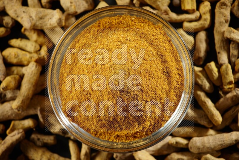 A glass bowl full of turmeric powder and dried whole fingers beside it on black flooring