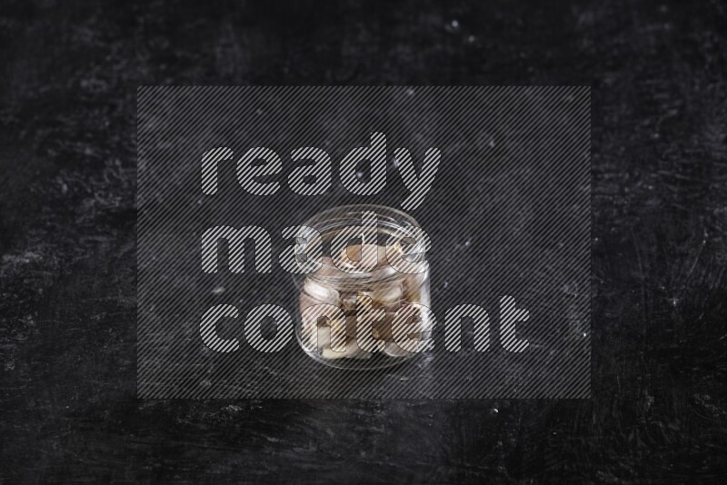 A glass jar full of garlic cloves on a textured black flooring in different angles