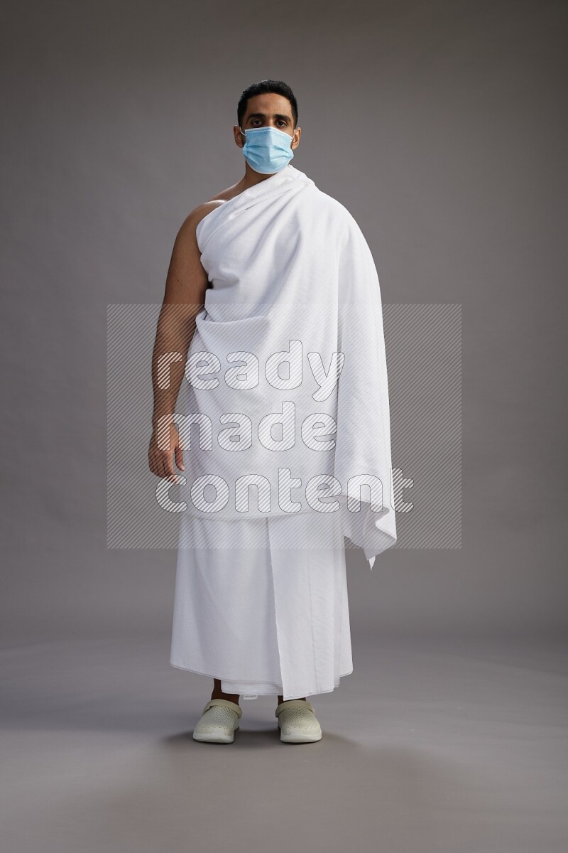A man wearing Ehram with face mask Standing Interacting with the camera on gray background