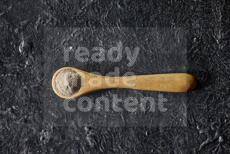 A wooden spoon full of white pepper powder on textured black flooring