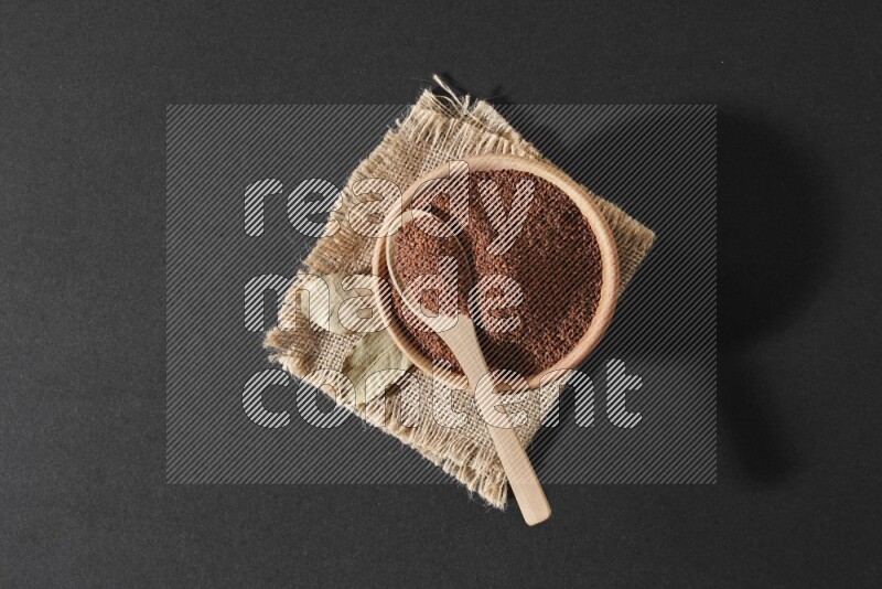 A wooden bowl full of garden cress seeds with wooden spoon full of the seeds on it on burlap fabric on a black flooring