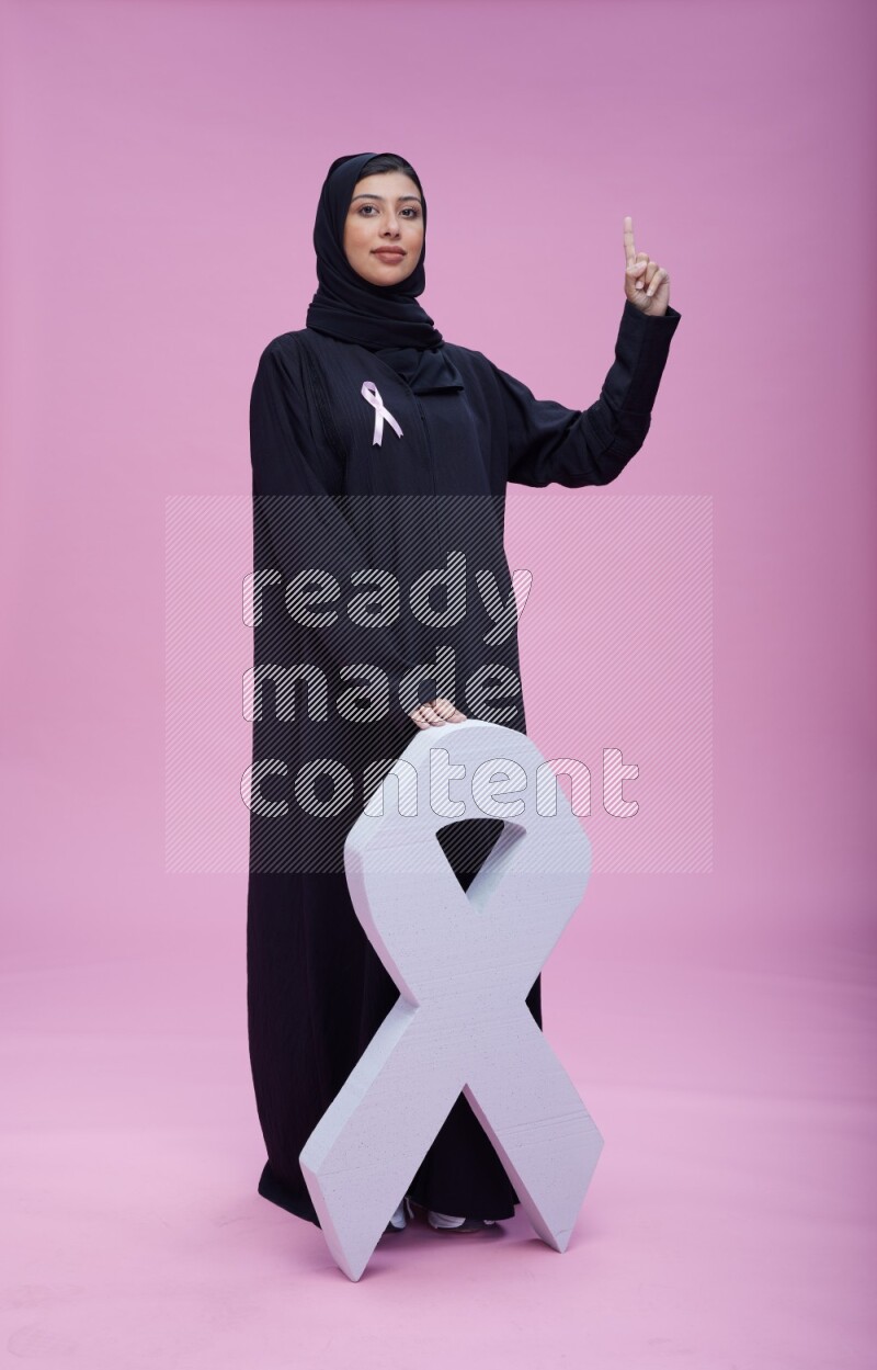 Saudi woman wearing pink ribbon on abaya standing holding awareness ribbon on plain pink background