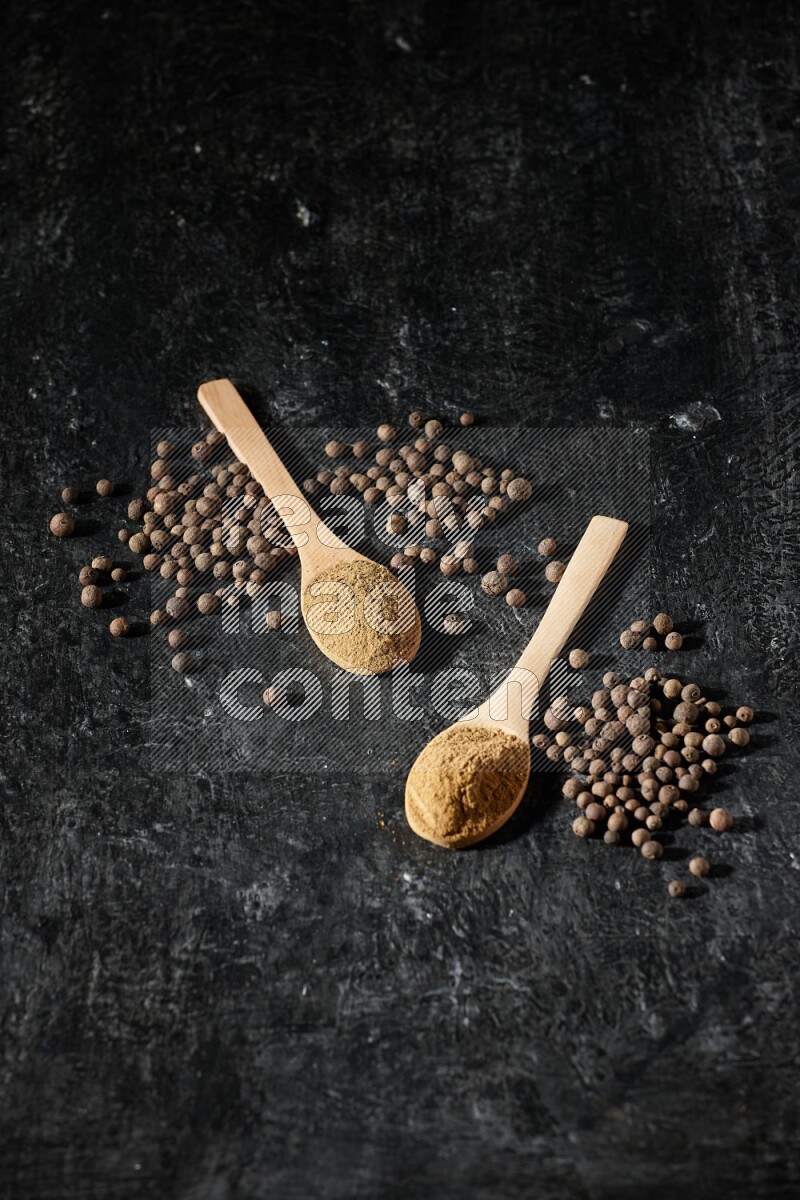Wooden spoons full of all spice powder and allspice whole balls beside it on a textured black flooring