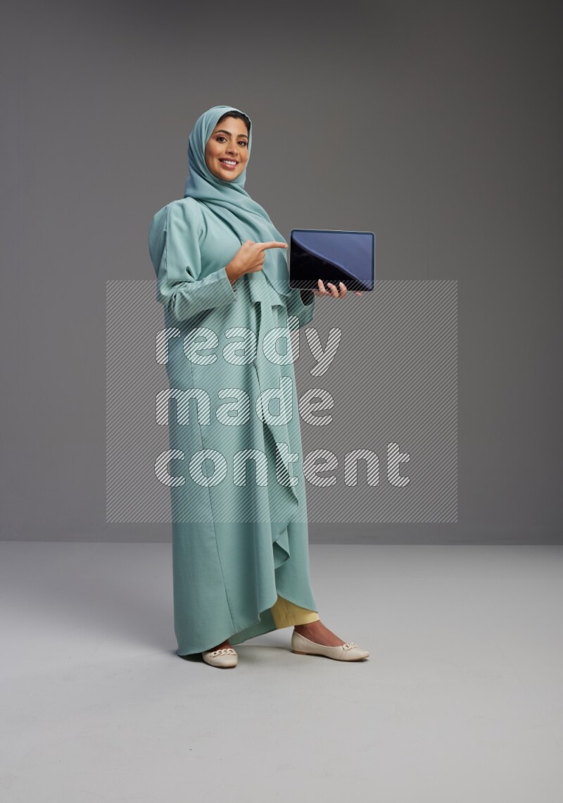 Saudi Woman wearing Abaya standing showing tablet to camera on Gray background