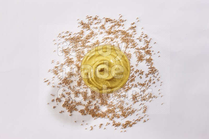 A glass bowl full of mustard paste with mustard seeds underneath on white flooring