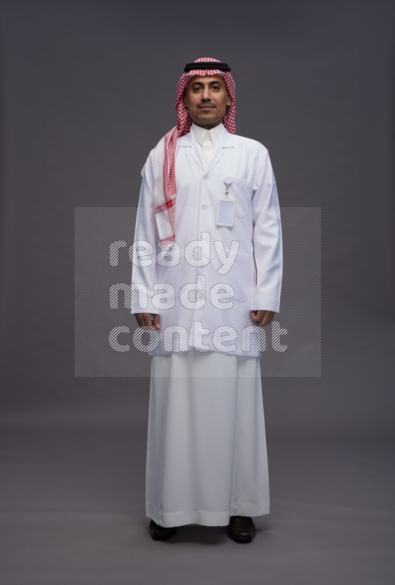 Saudi man wearing thob with lab coat and shomag with pocket employee badge standing interacting with the camera on gray background