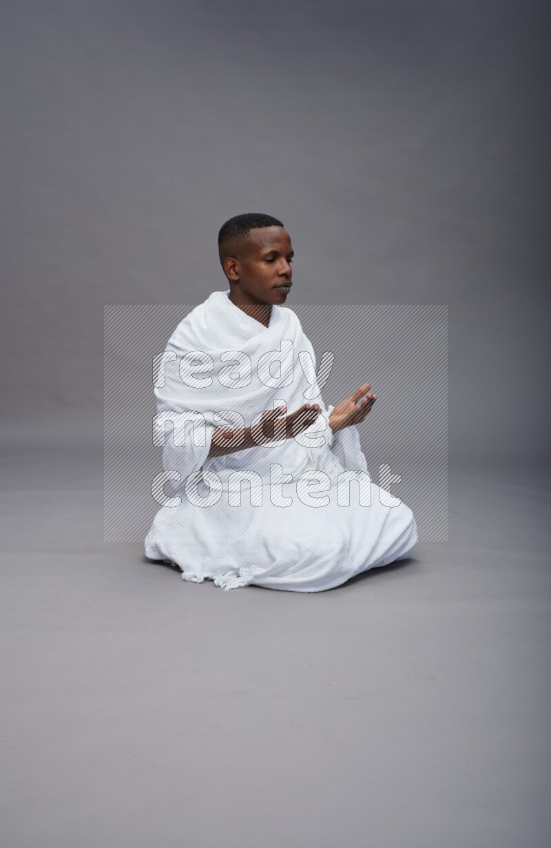 A man wearing Ehram sitting on floor dua'a on gray background