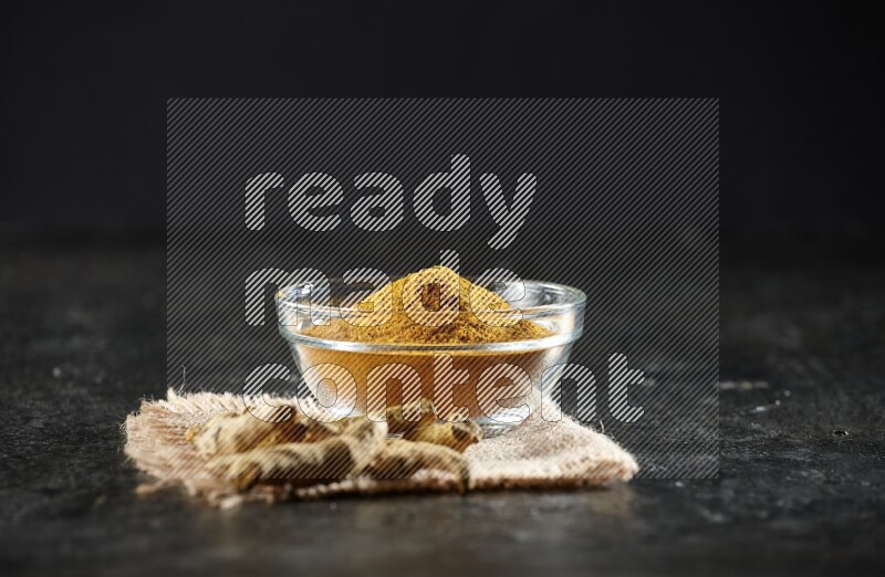 A glass bowl full of turmeric powder with dried turmeric fingers on a burlap fabric on textured black flooring