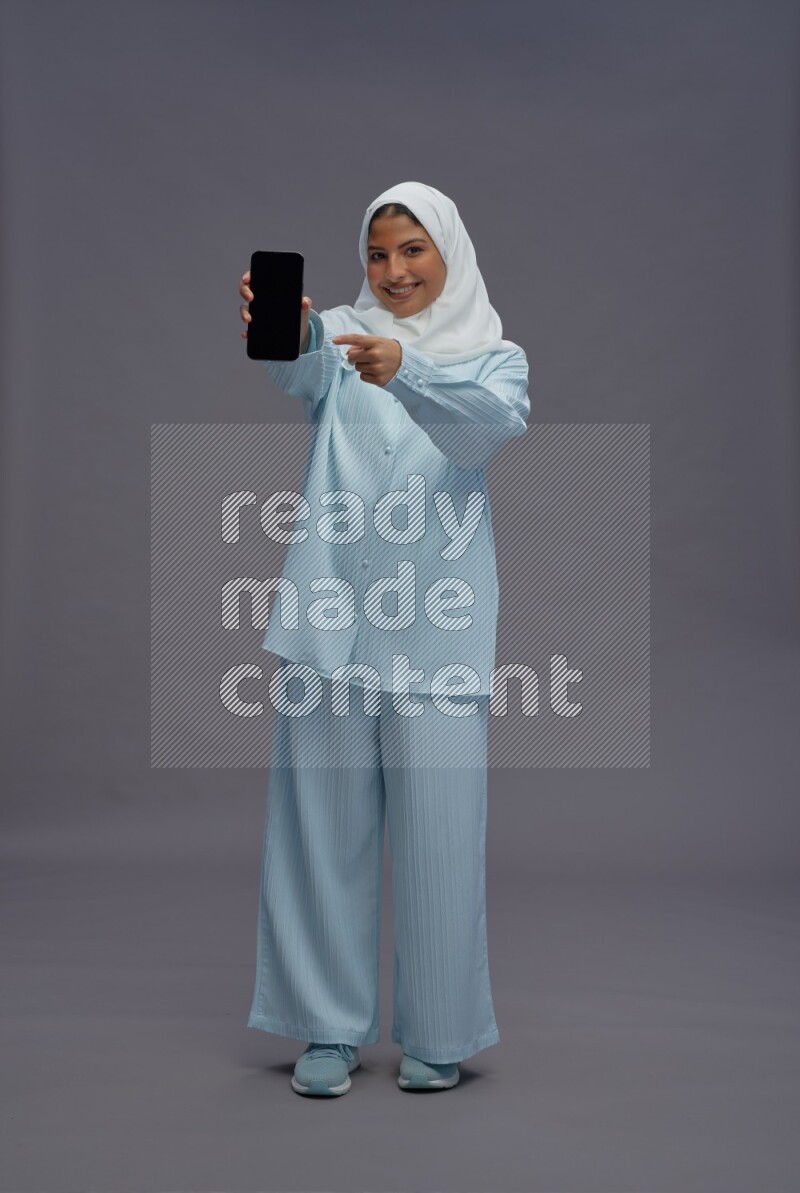 Saudi woman wearing hijab clothes standing showing phone to camera on gray background