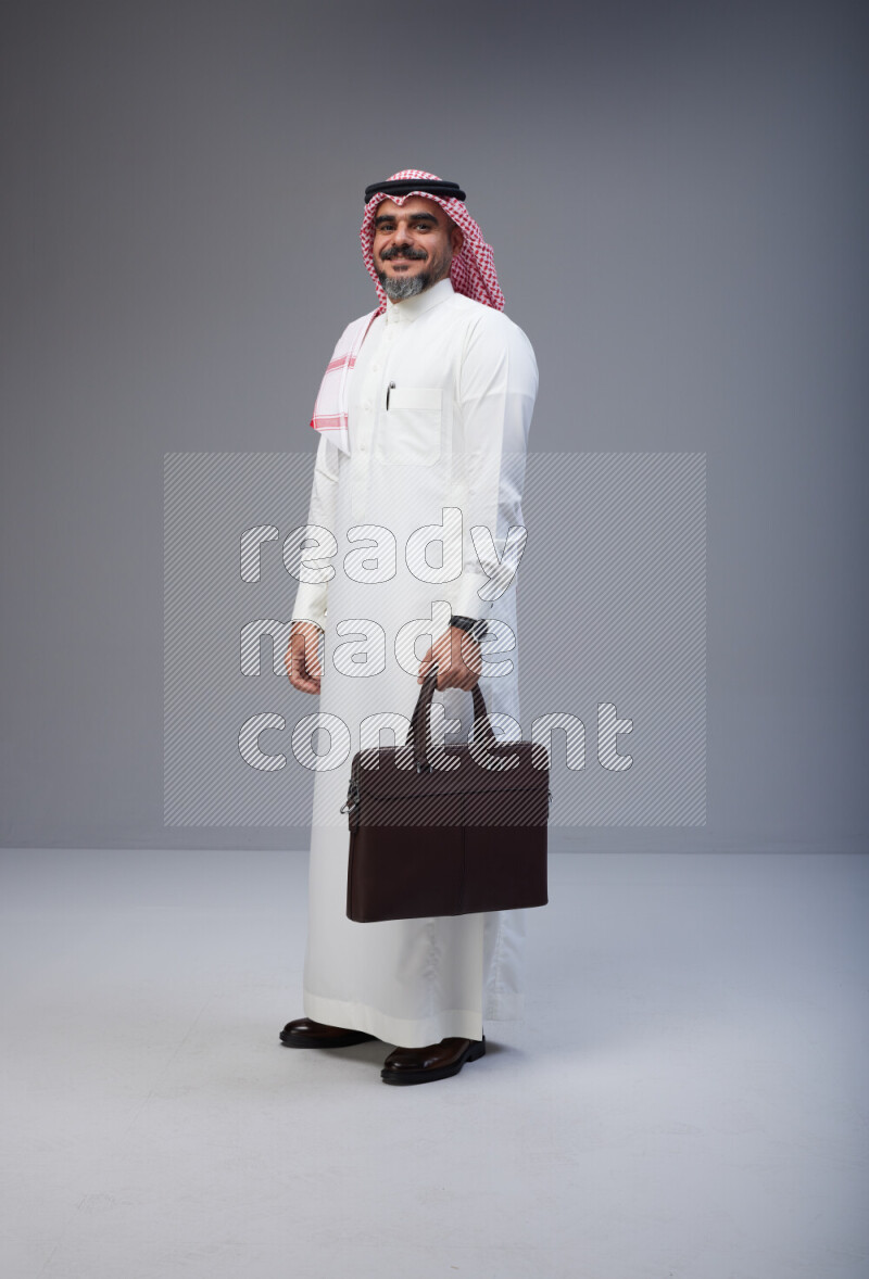 Saudi man Wearing Thob and red Shomag standing holding bag on Gray background