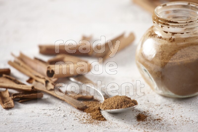 Herbal glass jar full cinnamon powder and a metal spoon surrounded by cinnamon sticks on a white background
