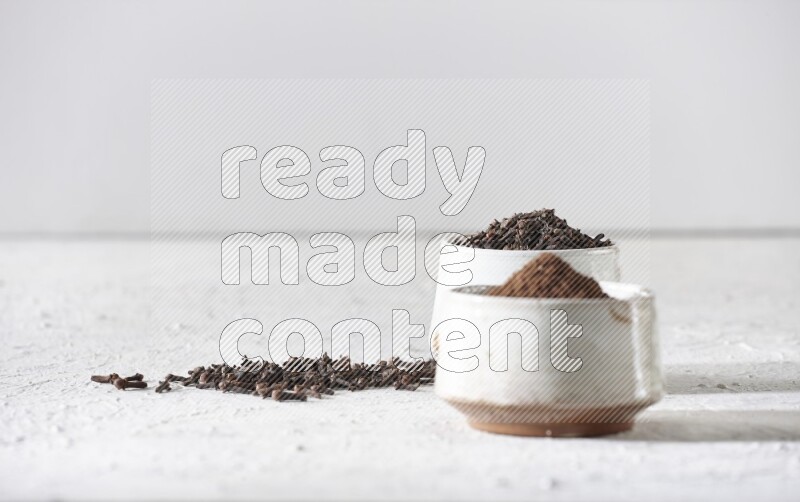 2 Beige ceramic bowls, one full of cloves and the other full of powder on textured white flooring