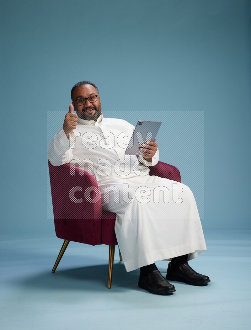 Saudi Man without shimag sitting on chair working on tablet on blue background