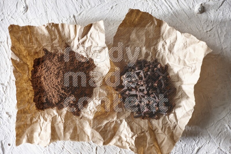 Cloves powder and cloves on 2 crumpled pieces of paper on a textured white flooring