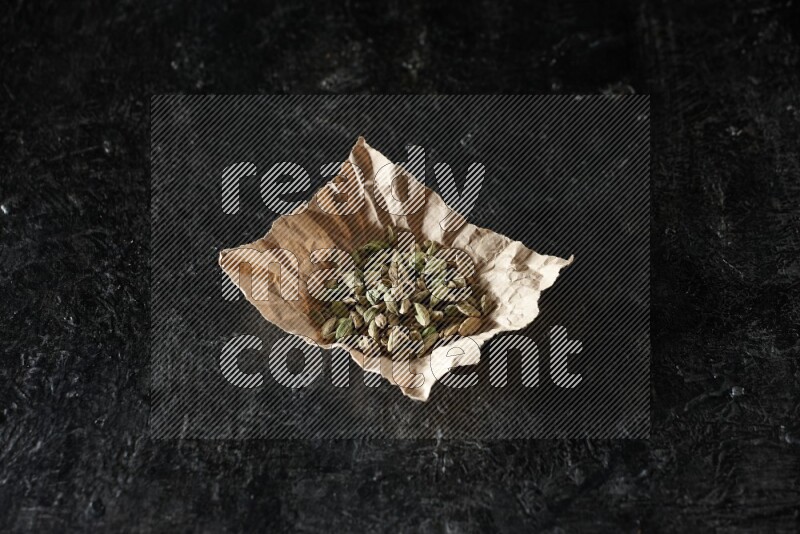 Cardamom seeds in a crumpled piece of paper on textured black flooring