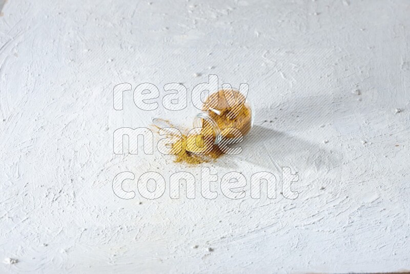 A flipped glass spice jar and a metal spoon full of turmeric powder and powder spilled out of it on textured white flooring