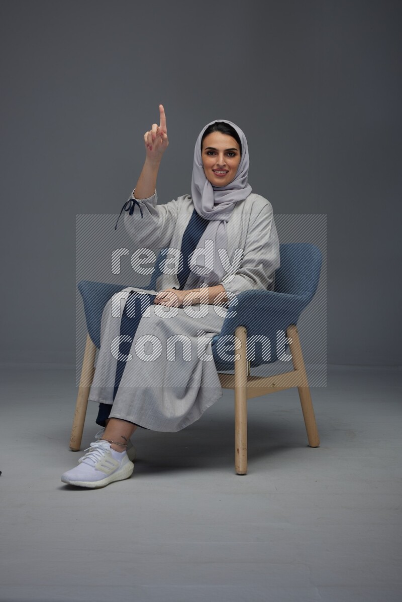 A Saudi woman wearing a light gray Abaya and head scarf sitting on a dark grey chair while making multi hand gestures eye level on a grey background